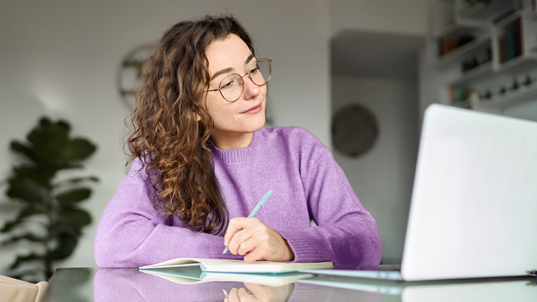 Eine Frau sitzt am Tisch, schreibt in ein Notizbuch und blickt dabei auf ihren Laptop.