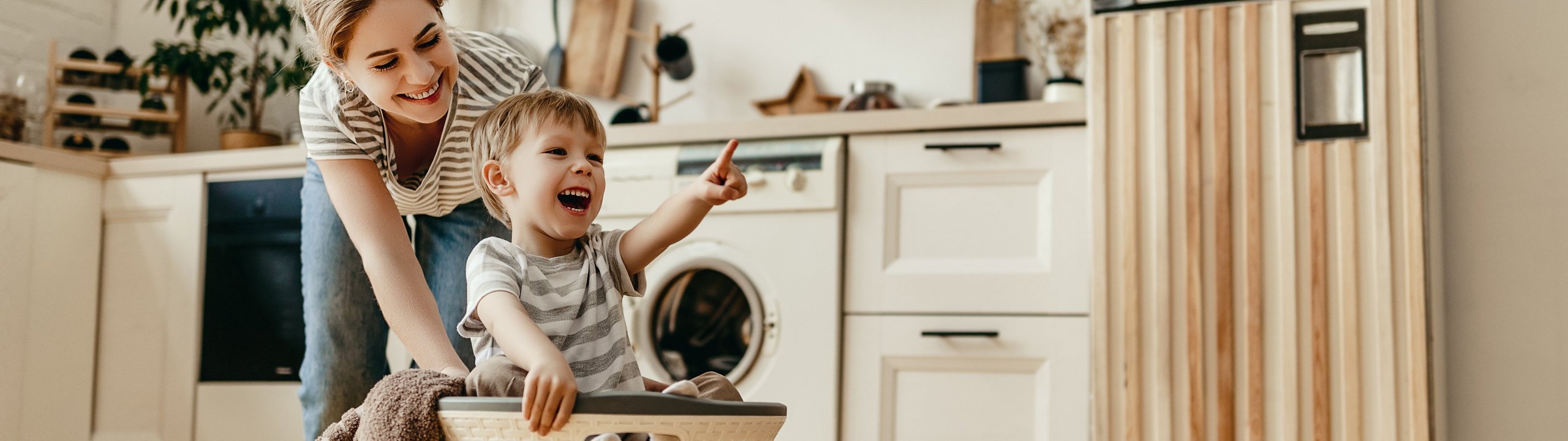 Mutter schiebt lachendes Kind im Wäschekorb durch die Küche