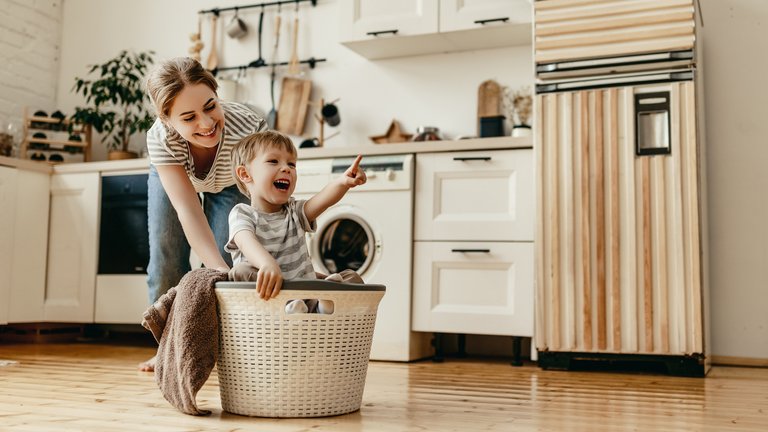 Mutter schiebt lachendes Kind im Wäschekorb durch die Küche