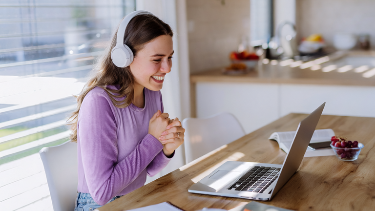 Eine junge Frau mit Kopfhörern lächelt vor ihrem Laptop bei einem Online-Meeting.