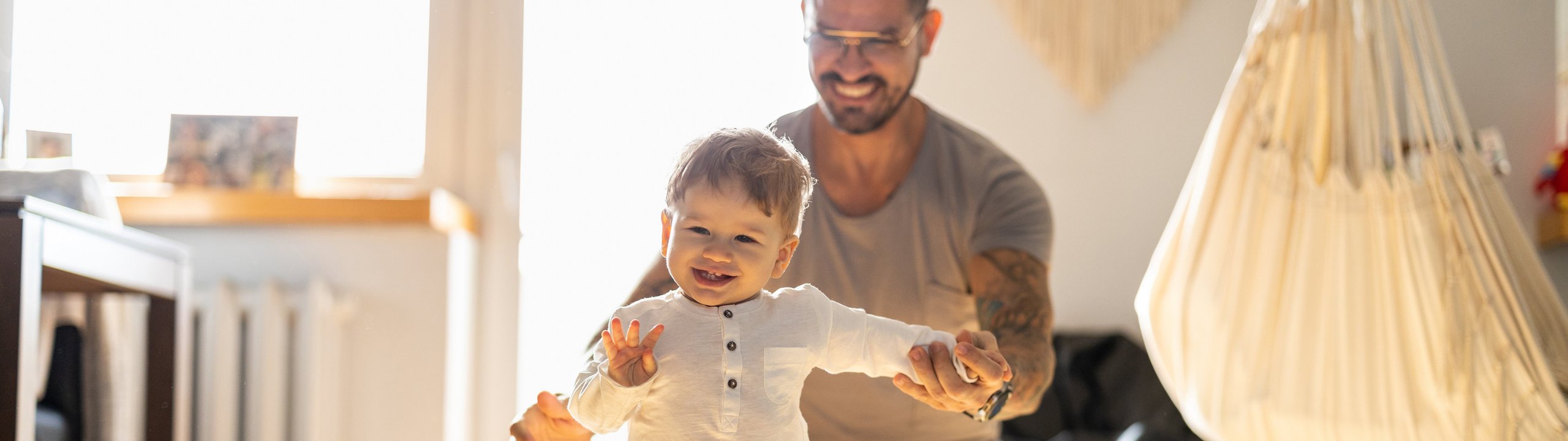 Vater unterstützt lachendes Kleinkind beim Laufenlernen im Wohnzimmer.