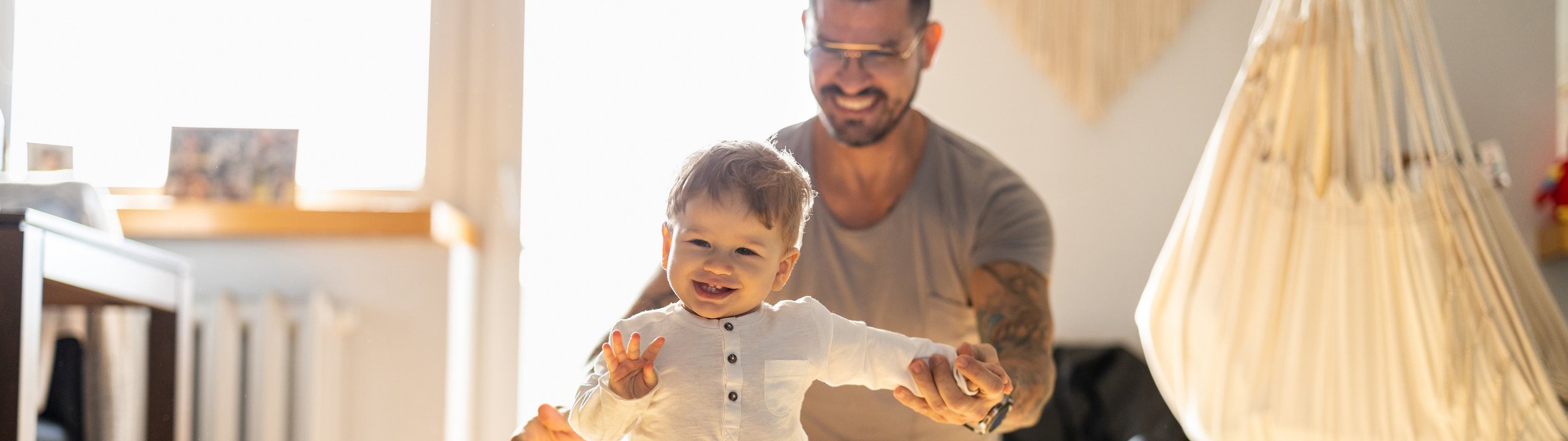 Vater unterstützt lachendes Kleinkind beim Laufenlernen im Wohnzimmer.