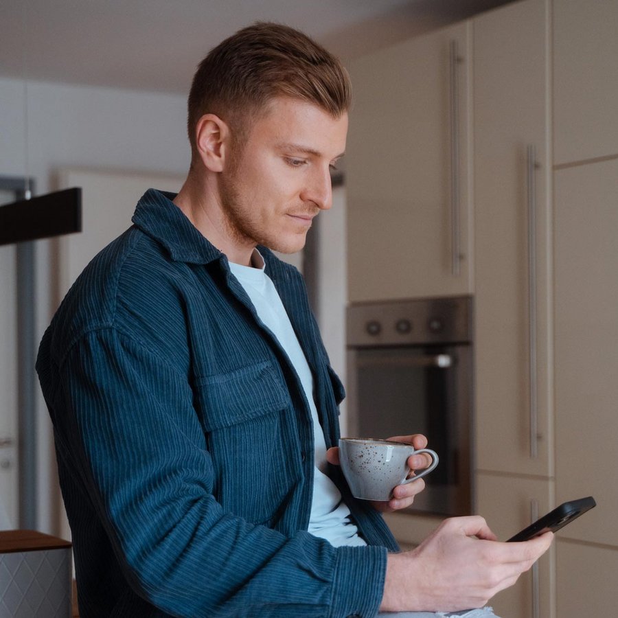 Mann sitzt mit Kaffee und Smartphone in der Küche.