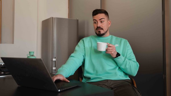 Ein Mann sitzt, mit einer Tasse in der Hand, erschrocken vor dem Laptop.