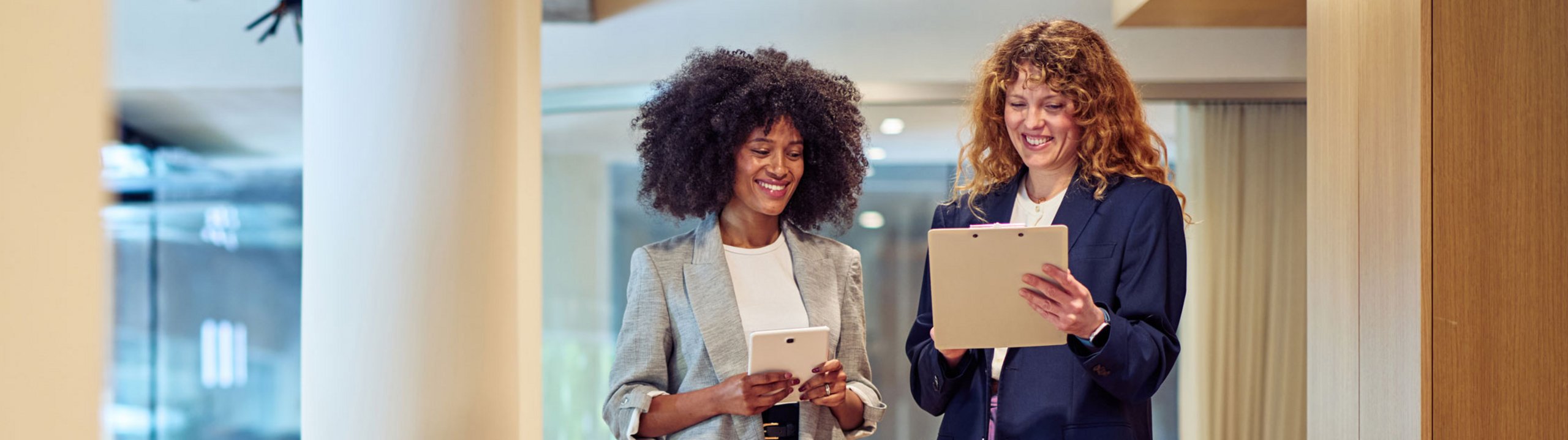 Zwei Frauen gehen gemeinsam durch ein Büro und schauen auf ein Tablet.