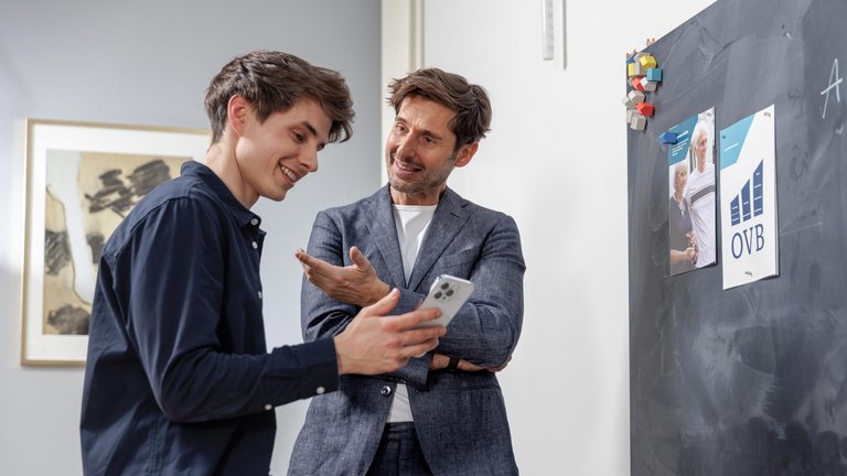 Zwei Männer besprechen etwas am Smartphone vor einer Tafel im Büro.