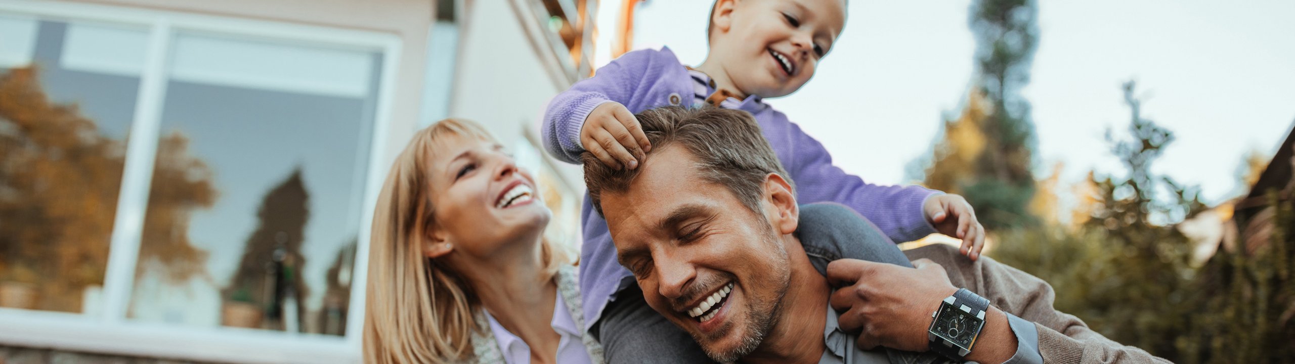 Familie spielt lachend im Garten, Kind sitzt auf den Schultern des Vaters.