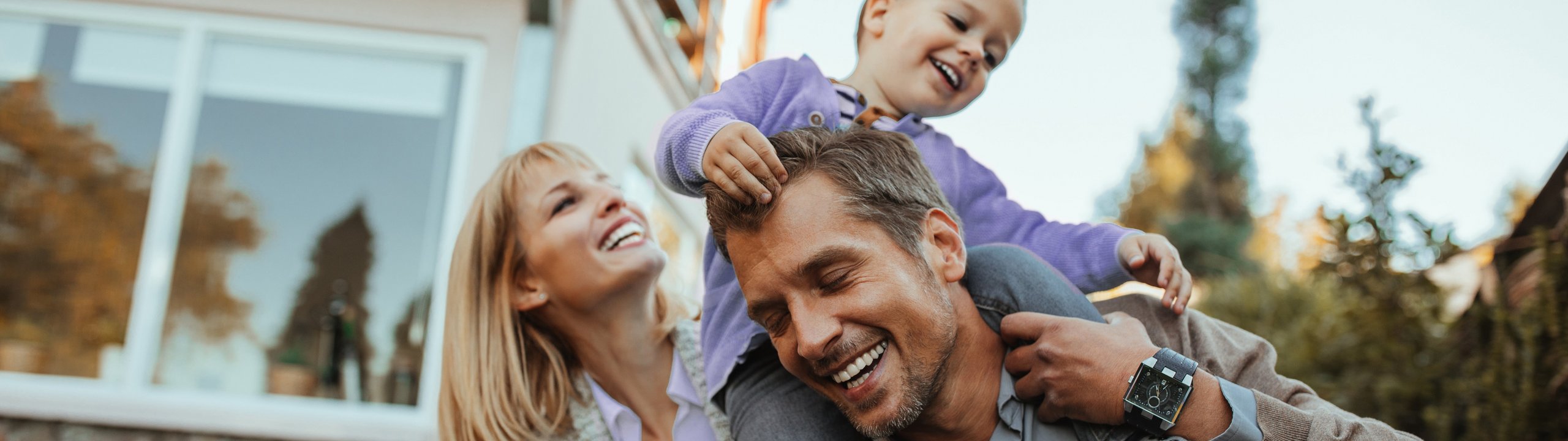 Familie spielt lachend im Garten, Kind sitzt auf den Schultern des Vaters.