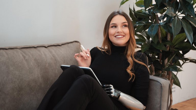 Frau mit Armprothese sitzt auf dem Sofa und schreibt auf einem Tablet.