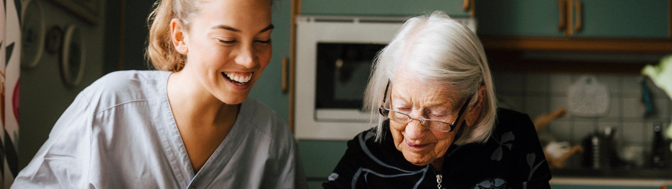 Pflegerin hilft älterer Frau beim Kreuzworträtzel am Küchentisch.