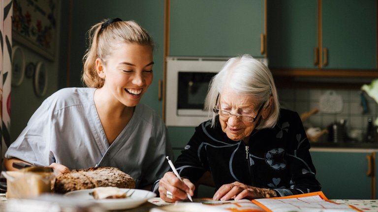 Pflegerin hilft älterer Frau beim Kreuzworträtzel am Küchentisch.