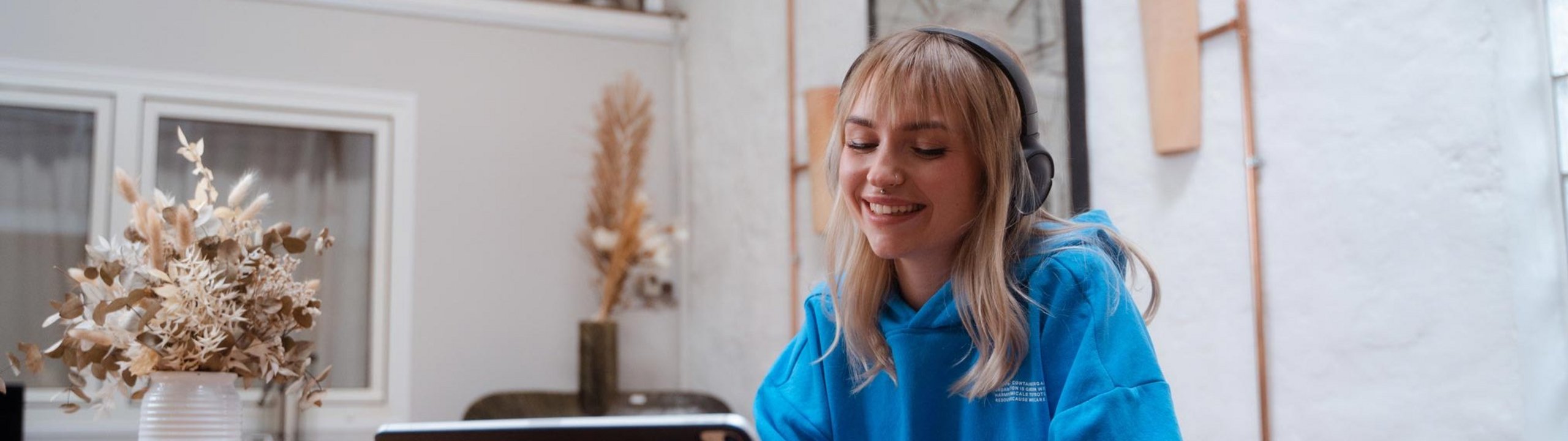 Junge Frau mit Kopfhörern arbeitet lächelnd an einem Tablet am Tisch.
