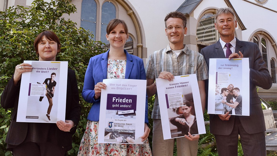 Vier Menschen halten vor Kirche Poster des Projekts „Frieden geht anders“ hoch