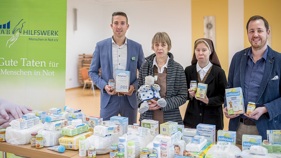 4 Personen sammeln Verbrauchsmaterial für bedürftige Familien mit Säuglingen. Die Spenden liegen vor den Personen aufgereiht auf einem Tisch