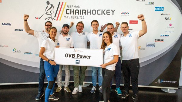 Jens Karsten und sein OVB Team stehen vor einem großen Plakat der German Chairhockey Masters 2019 und halten ein Schild hoch auf dem OVB Power steht. Alle gucken freudig und zwei Männer recken die Fäuste hoch