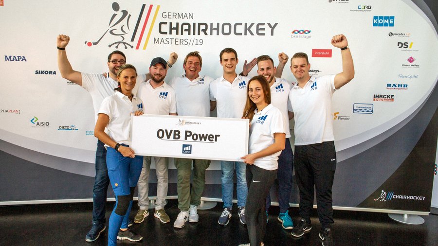 Jens Karsten und sein OVB Team stehen vor einem großen Plakat der German Chairhockey Masters 2019 und halten ein Schild hoch auf dem OVB Power steht. Alle gucken freudig und zwei Männer recken die Fäuste hoch