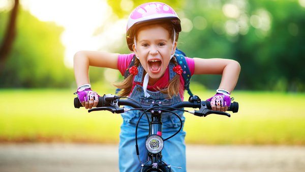 Mädchen mit rosa Fahrradhelm und Latzhose fährt Fahrrad, beugt sich über den Lenker und schreit freudig. Es ist Sommer. Der Hintergrund ist unscharf