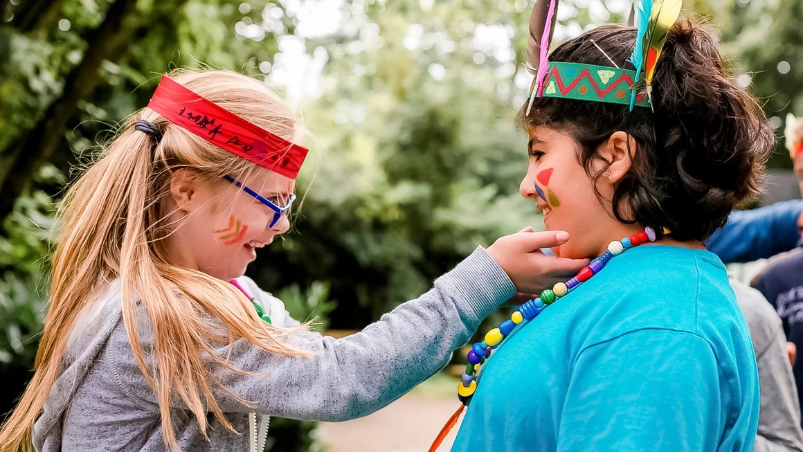 Zwei Mädchen sind als Indianerinnen verkleidet. Rechtes Mädchen packt linkem Mädchen liebevoll an die Wange