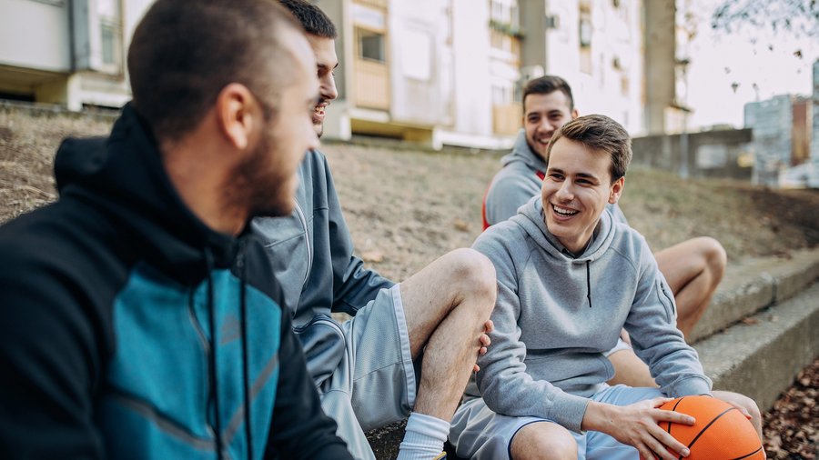 Vier Männer sitzen auf Treppenstufen und erzählen sich etwas. Dabei lachen sie. Sie tragen Kaputzenpullover da sie eine Pause vom Basketballspielen machen