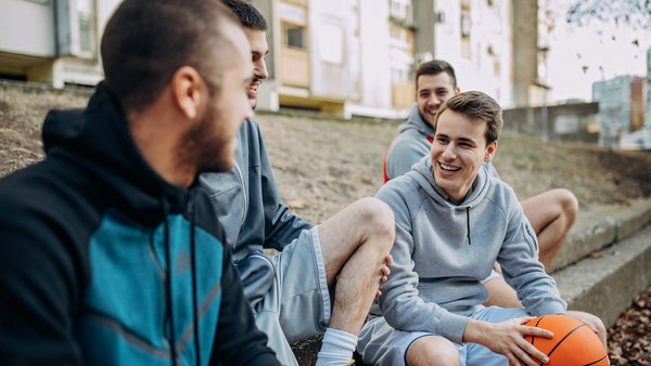 Vier Männer sitzen auf Treppenstufen und erzählen sich etwas. Dabei lachen sie. Sie tragen Kaputzenpullover da sie eine Pause vom Basketballspielen machen