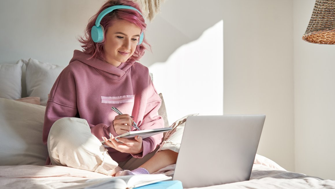 Jugendliches Mädchen sitzt auf Bett mit Computer und Kopfhörern