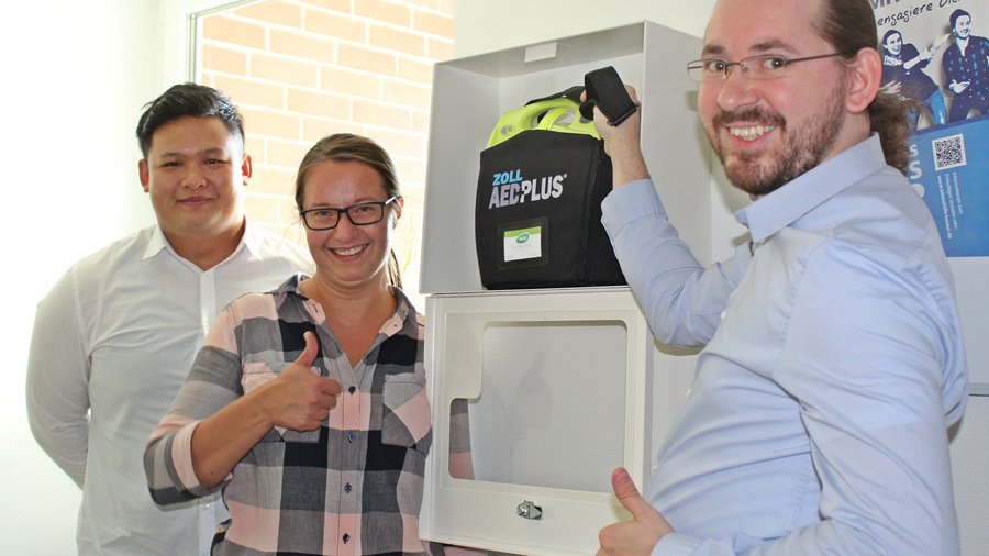 Zwei Männer und eine Frau stehen vor einem AED Defibrillator
