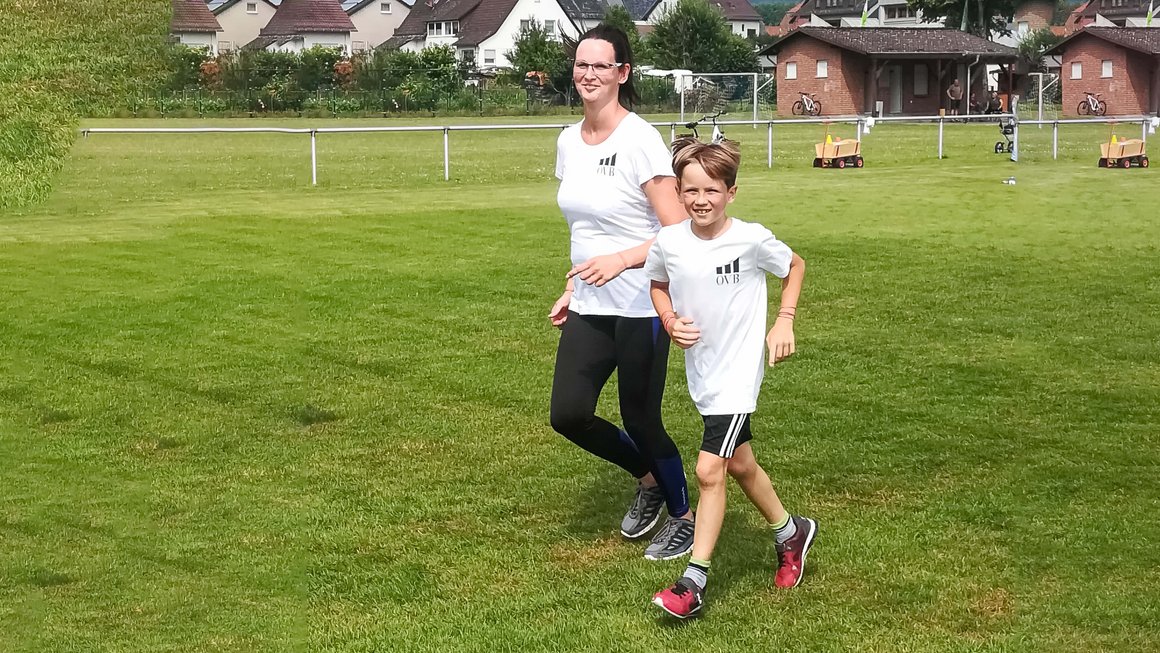 Frau und Kind laufen auf Wiese und blicken fröhlich in die Kamera bei Sommerwetter