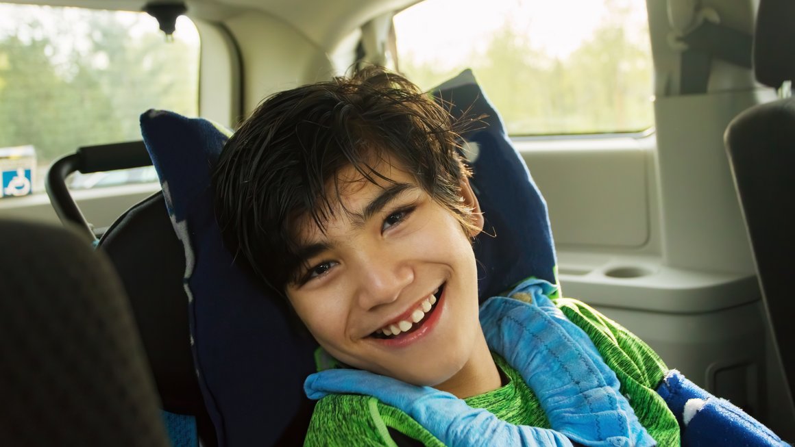 Behinderter Junge mit dunklen Haaren sitzt im Kindersitz in einem Auto und lächelt glücklich