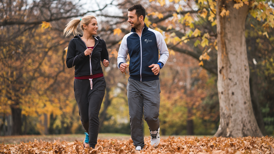 Mann und Frau sind mittig im Bild und schauen sich beim Laufen liebevoll in die Augen. Im Hintergrund ist ein herbstlicher Wald mit bräunlichen Blättern zu sehen