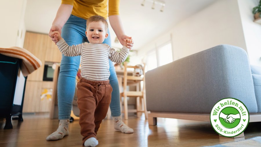 Kleiner Junge lernt laufen. Er wird an beiden Händchen festgehalten von einer Frau die nur bis zum Hals zu sehen ist