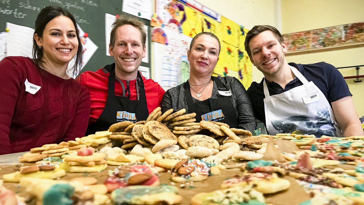 Tisch ist bedeckt mit Weihnachtsplätzchen. Hinter dem Tisch sitzen zwei Frauen und zwei Männer mit Schürzen und freuen sich