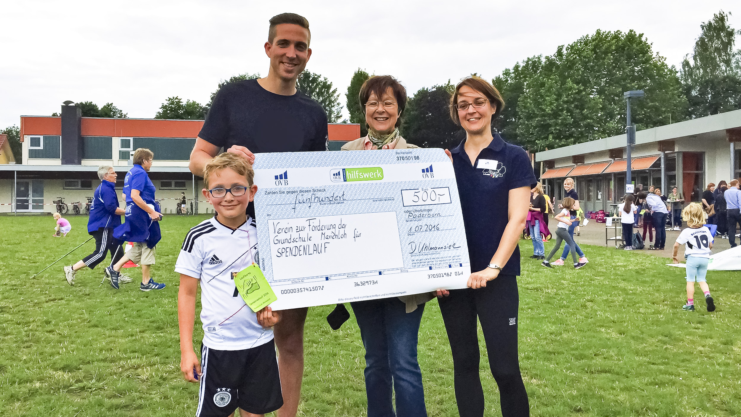Mann und Junge mit Brille übergeben Spendenscheck an Schulleiterin der Grundschule Marienloh beim Spendenlauf