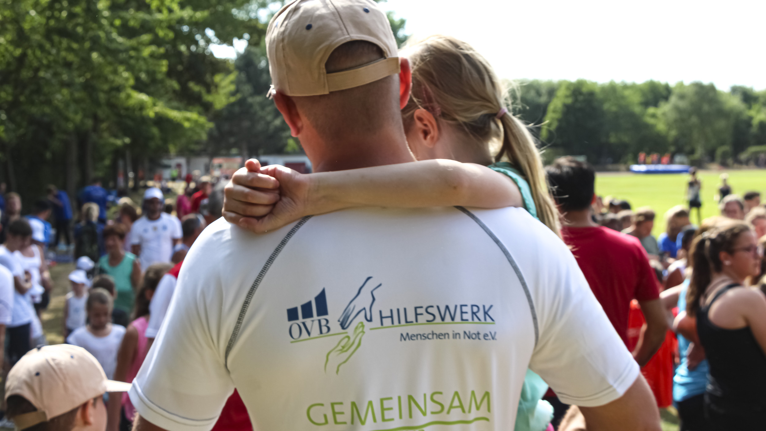 Mann in Großaufnahme geht draußen mit Rücken zugewandt und kleinem Mädchen auf dem Arm nach vorne. Unscharf sind links und rechts viele Leute zu sehen. Es ist Sommer