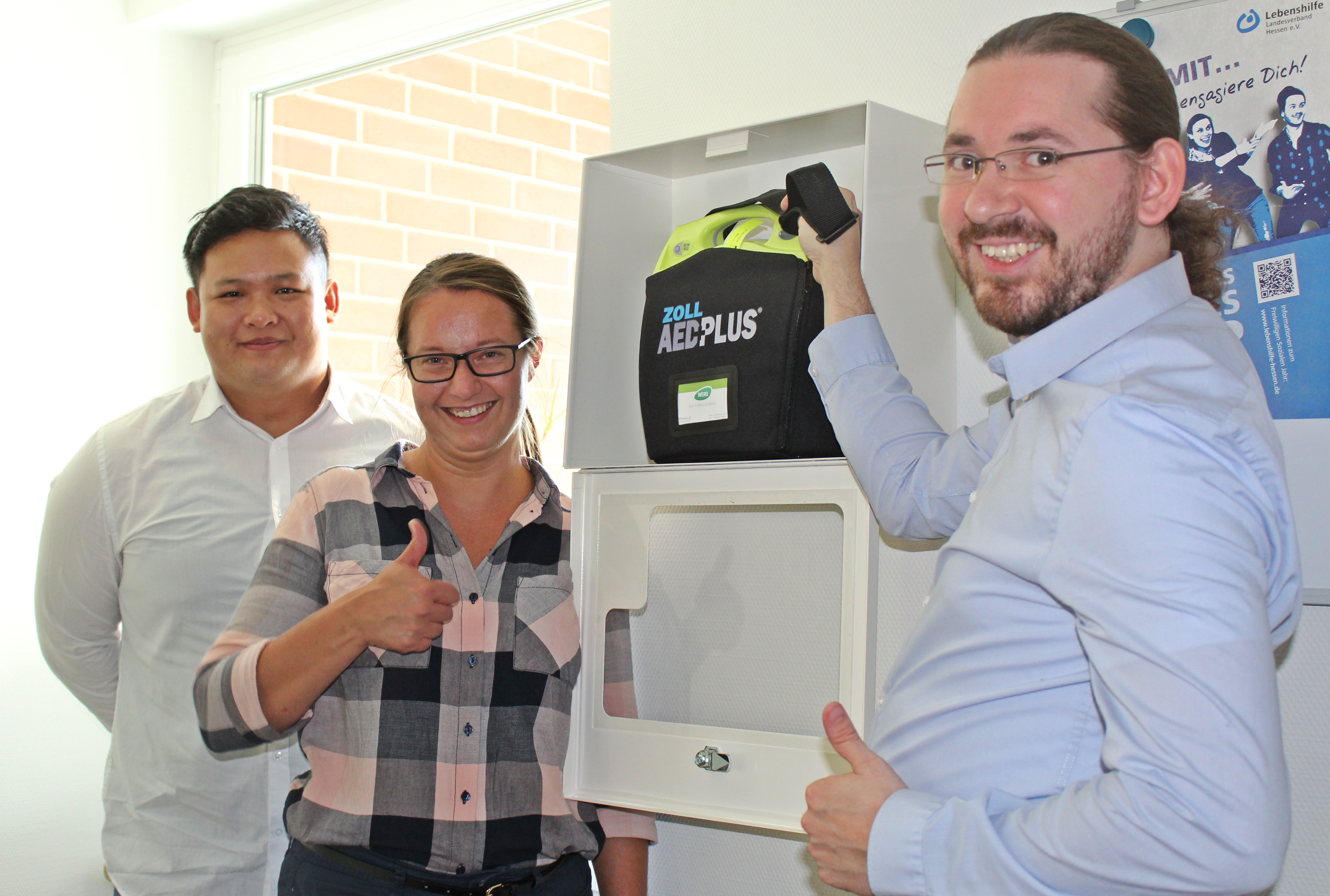 Zwei Männer und eine Frau stehen vor einem AED Defibrillator
