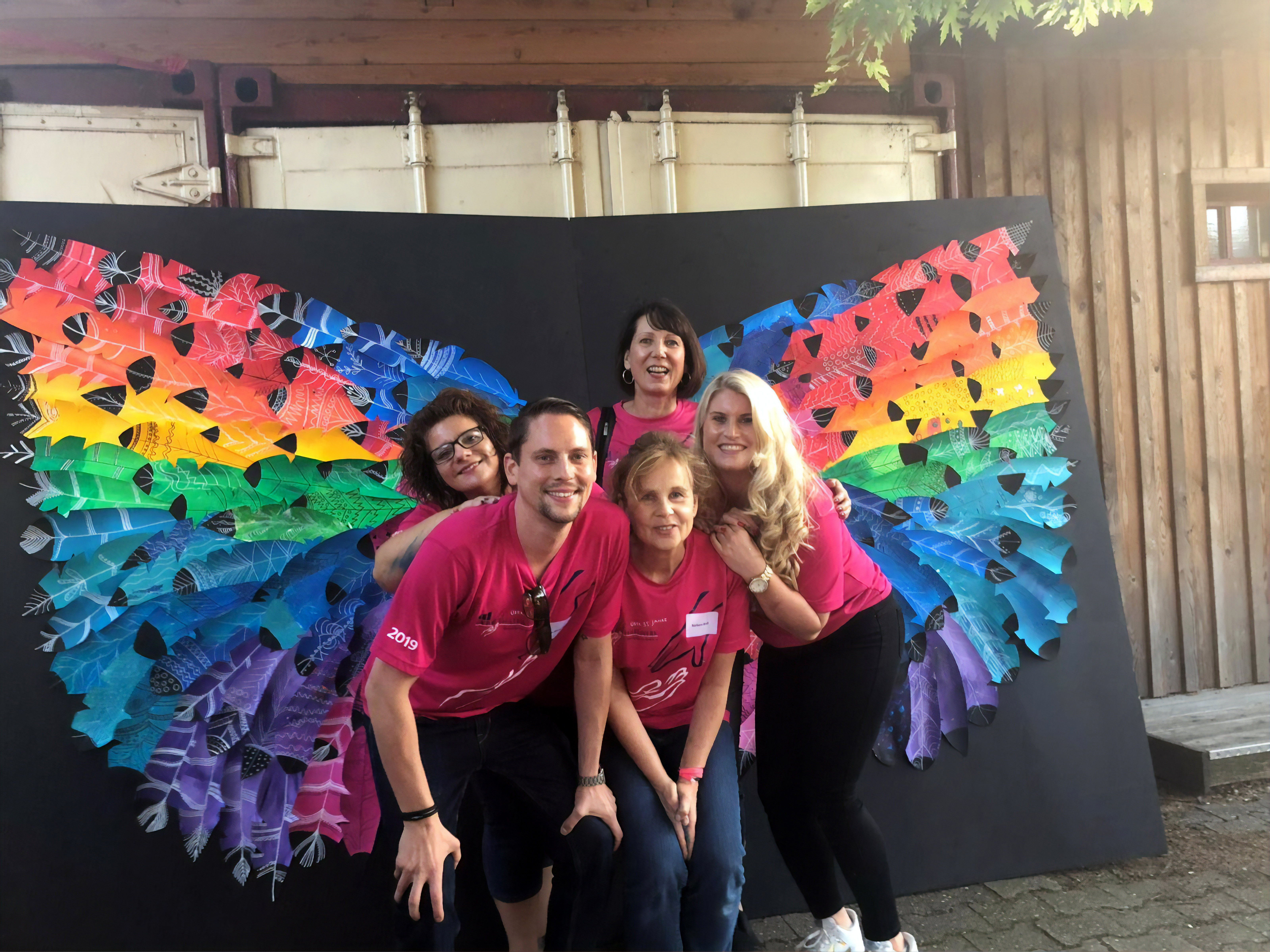 5 OVB Mitarbeiter mit rose Tshirts stehen vor schwarzem Hintergrund auf dem in regenbogenfarben Flügel aufgezeichnet sind