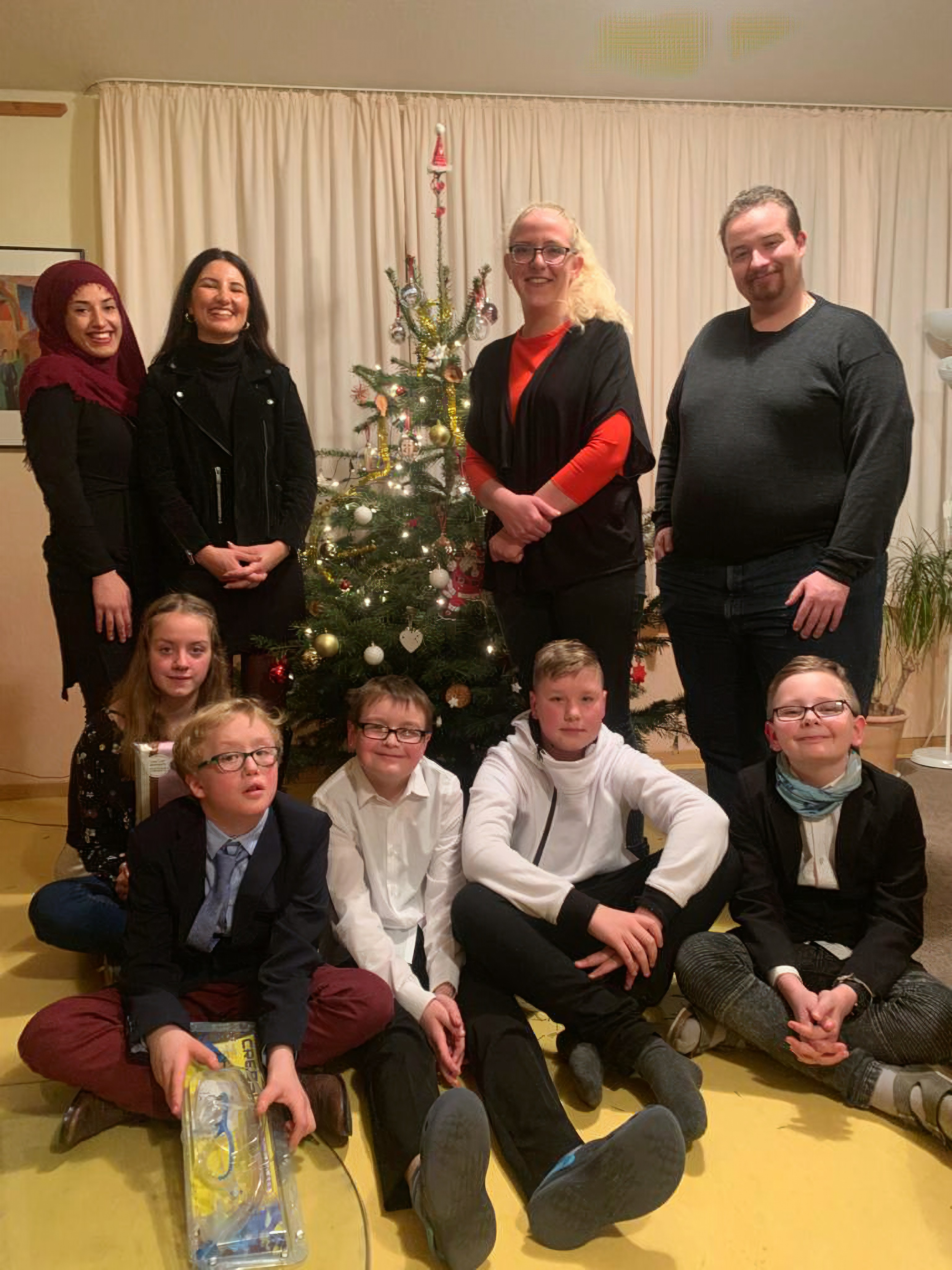 Mehrere Kinder sitzen auf dem Boden, 4 Erwachsene stehen dahinter. In der Mitte ist eine Weihnachtsbaum zu sehen