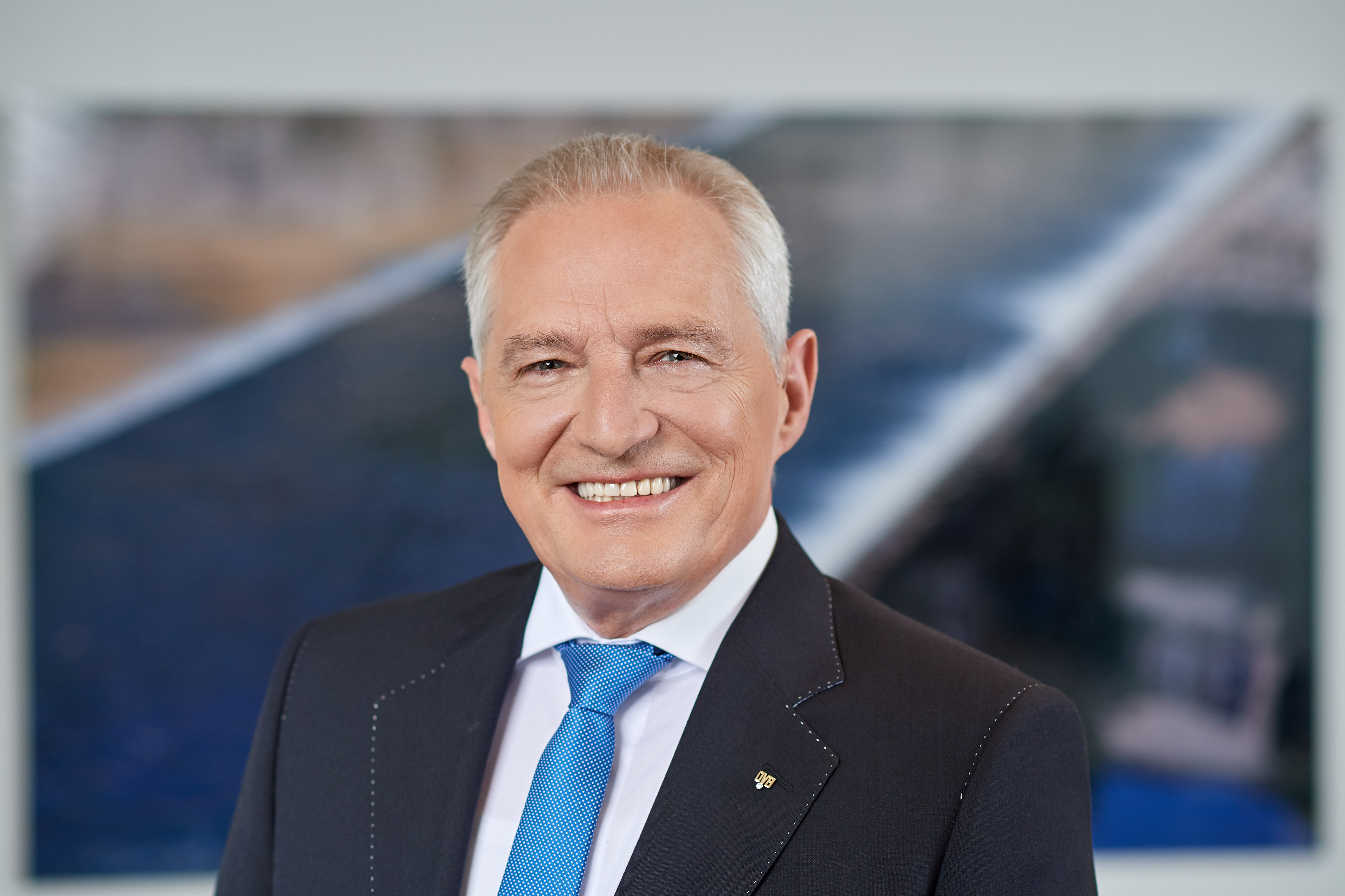 Oskar Heitz im dunklen Anzug mit blau gepunkteter Krawatte. Er lächelt und der Hintergrund ist unscharf