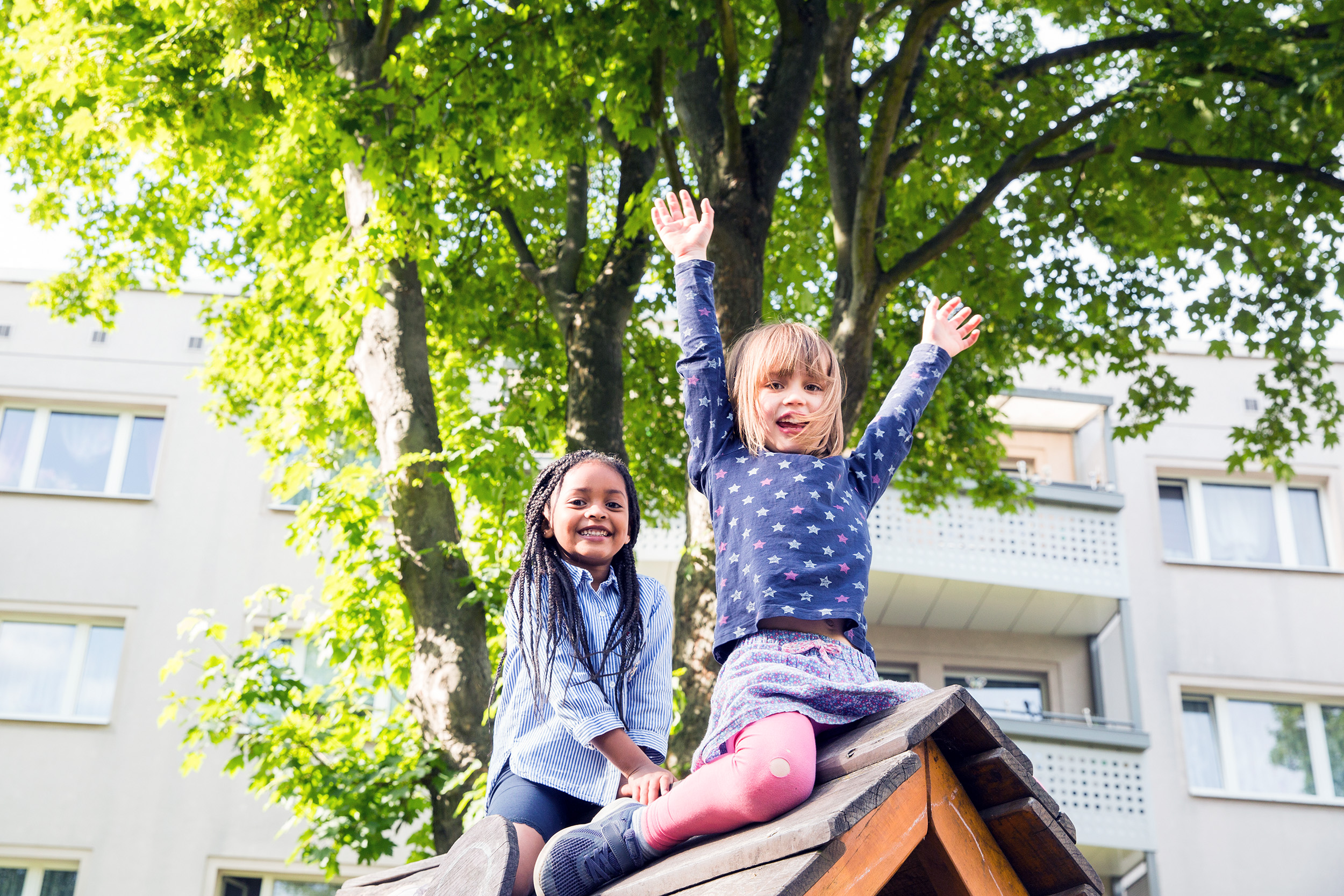 Zwei Mädchen sitzen auf Spielhaus eines Spielplatz und freuen sich im Sommerwetter