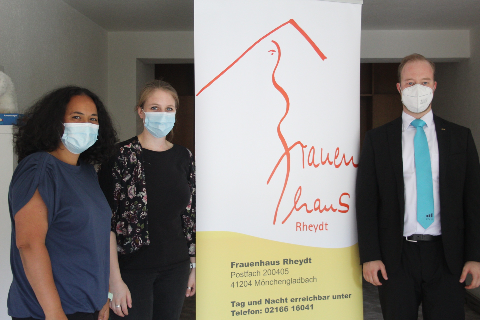 Zwei Frauen und ein Mann stehen mit Maske aufgereiht. In der Mitte ist ein Banner für die Spendensammelaktion des Frauenhaus in Mönchengladbach Rheydt